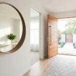 Round wooden mirror on white wall in bright entryway with open front door, reflecting green plant and natural light, enhancing Feng Shui.